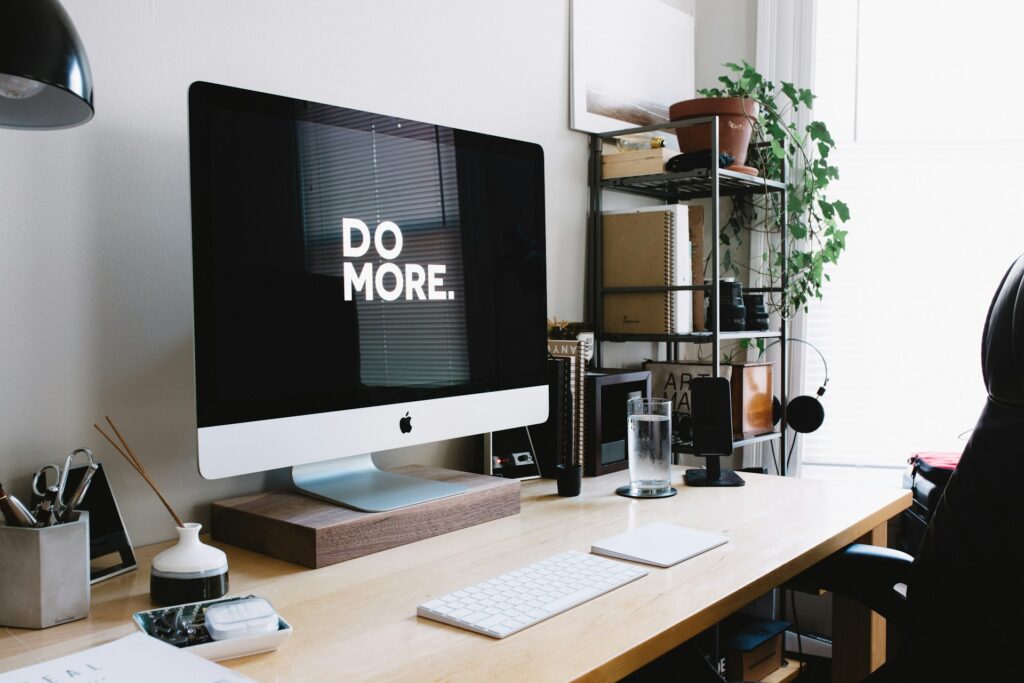 Minimalist home office desk with an iMac displaying “DO MORE.” surrounded by notebooks, plants, and workspace essentials.